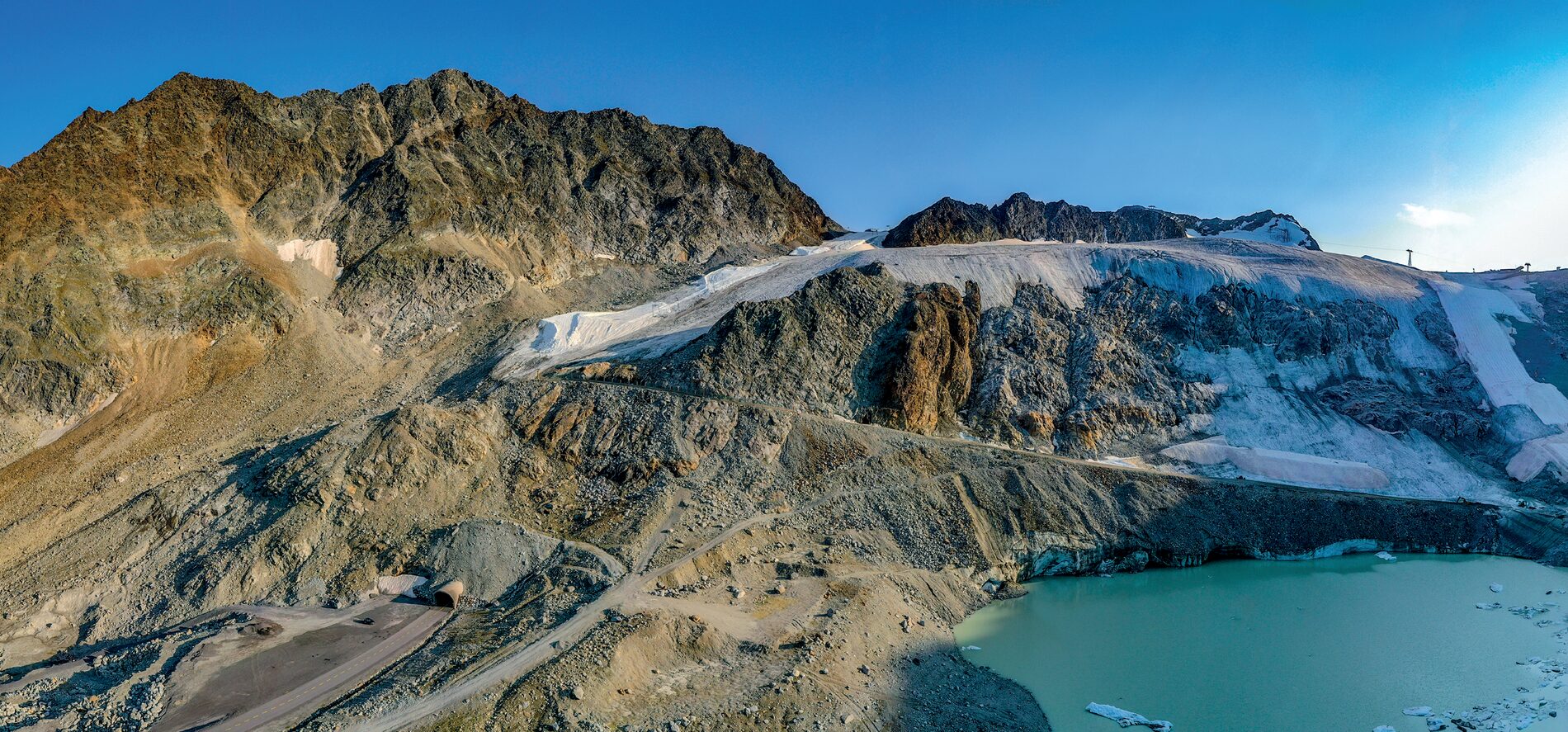 Gletscher über einem türkisfarbenen Schmelzwassersee, links ein Tunnelportal in einer Geröllhalde