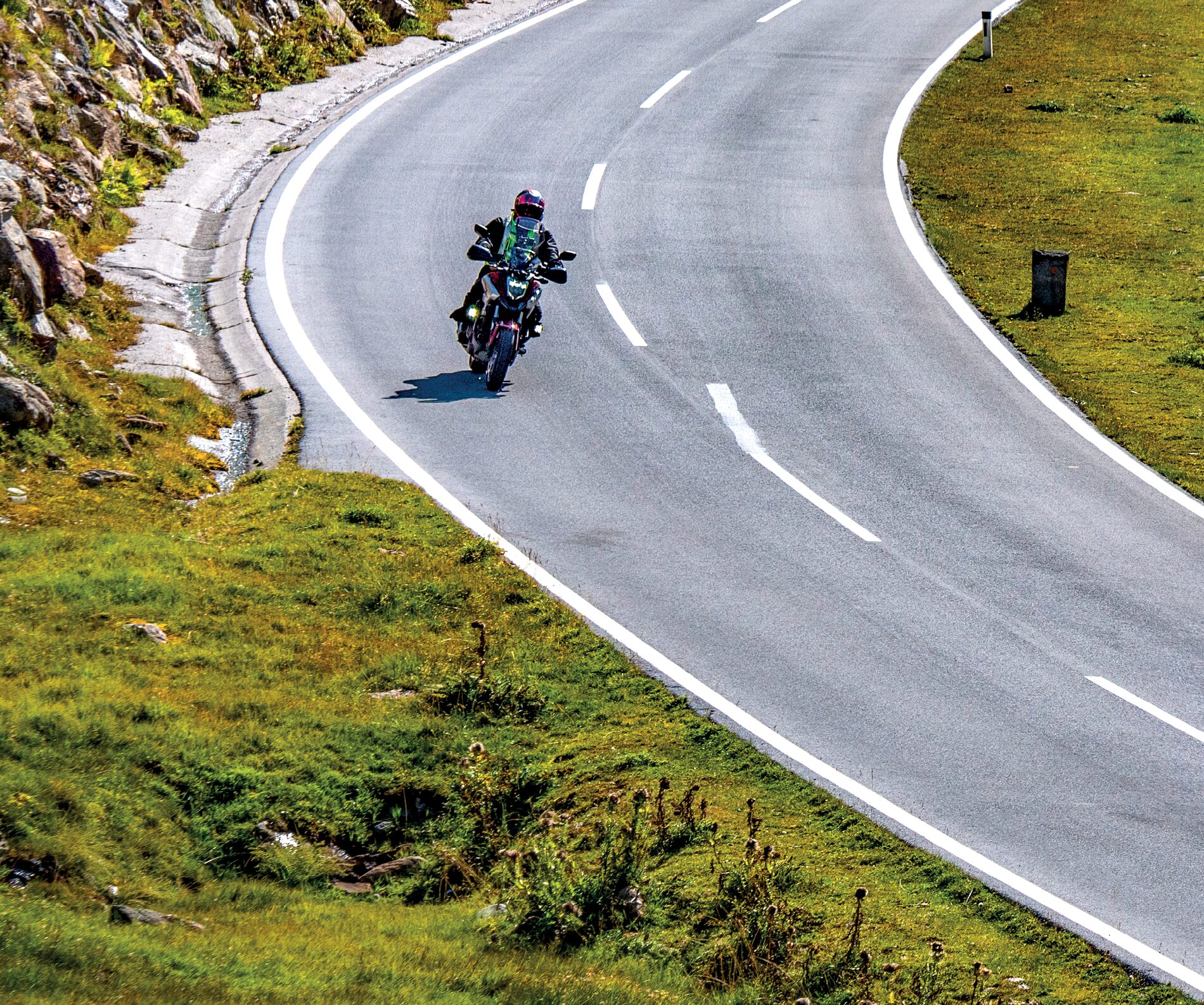 Motorradfahrer in einer weiten Kurve zwischen Grashang und Steinmauer