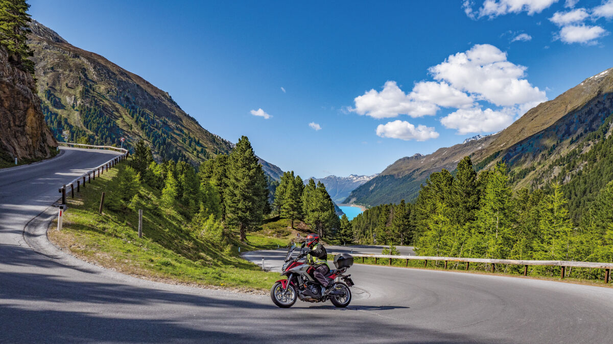 Motorrad in einer Linkskurve, im Tal ein türkisfarbener Stausee, rundum bewaldete Hänge und Gipfel