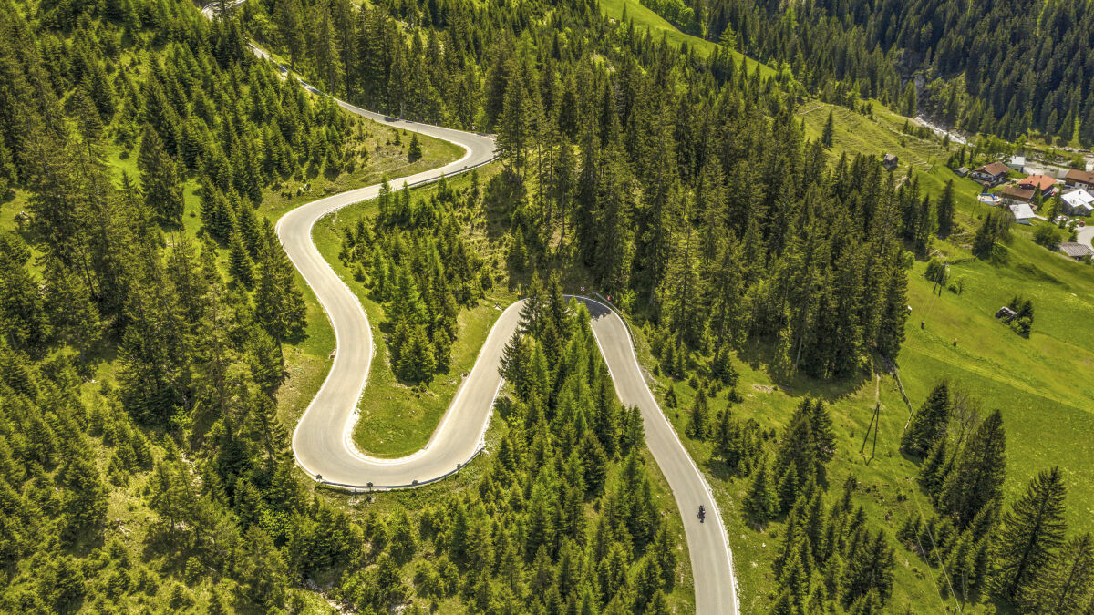 Luftaufnahme einer Bergstraße mit vier Kehren durch Fichtenwald, ein einzelnes Motorrad, rechts die Häuser eines Dorfs