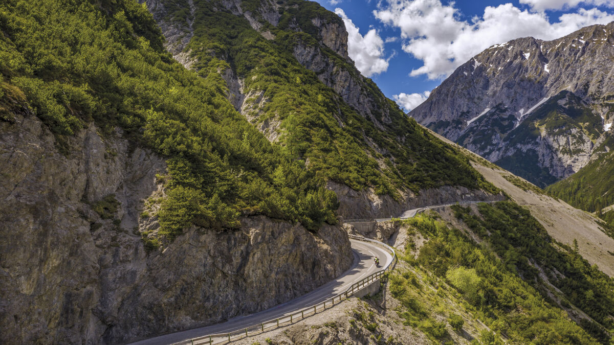 Zwei Motorräder in einer Kehre unter einer Felswand, gegenüber ein Kalkmassiv mit Schneeresten