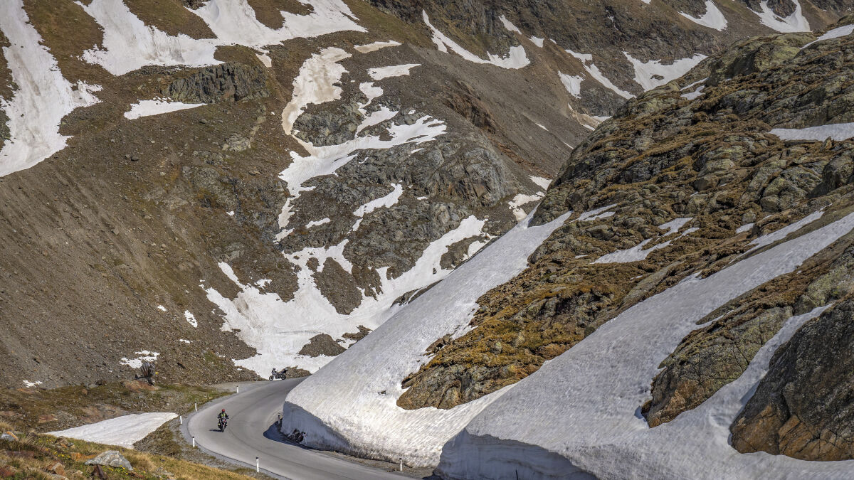 Zwei Motorräder in einer Haarnadelkurve, seitlich eine hohe Wand aus Altschnee, oben Felshänge mit Schneefeldern